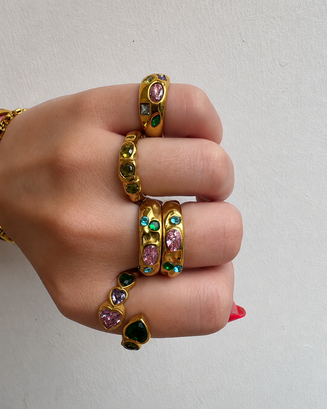 A hand wearing multiple gold rings and a bracelet, each set with colorful gemstones including pink, green, and dark blue, against a plain light gray background.
