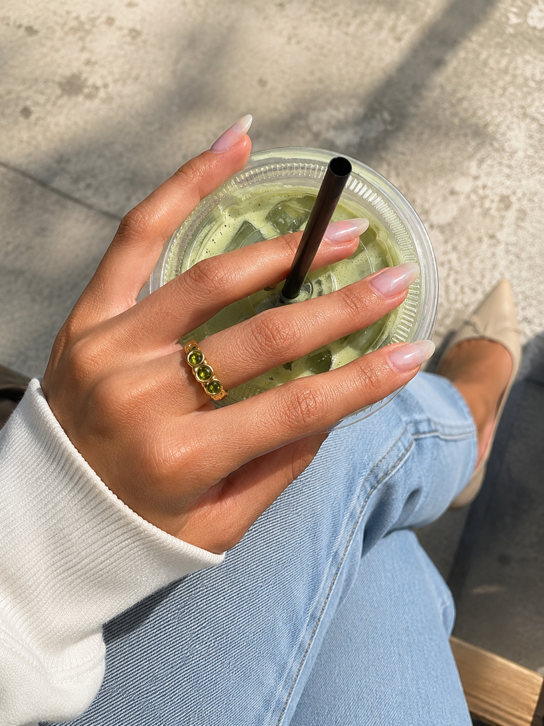 A close-up lifestyle shot of a hand holding an iced green drink with a black straw, wearing a gold ring with green stones. The person is dressed in a white long-sleeve top and light blue jeans, seated outdoors on a concrete surface.