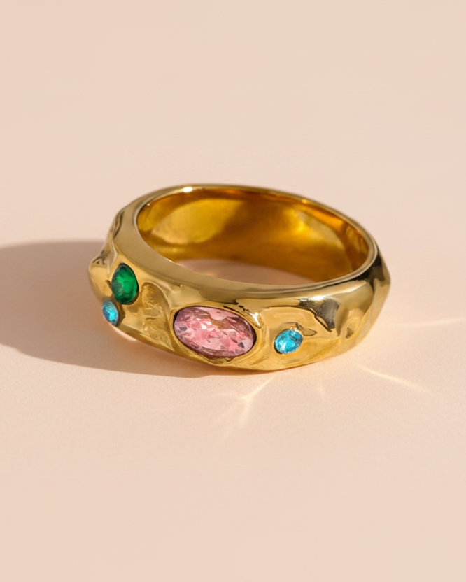 A gold ring with a wavy, organic textured band, featuring a central oval pink gemstone and smaller green and blue accent gemstones, set against a solid pale peach background.