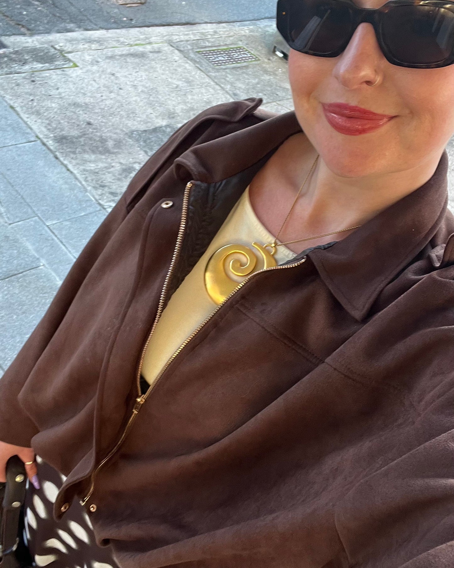 A woman wearing a brown suede-like zip-up jacket over a gold top with a spiral necklace, black sunglasses, and a black-and-white patterned skirt, photographed in a selfie-style outdoor shot on a paved sidewalk.