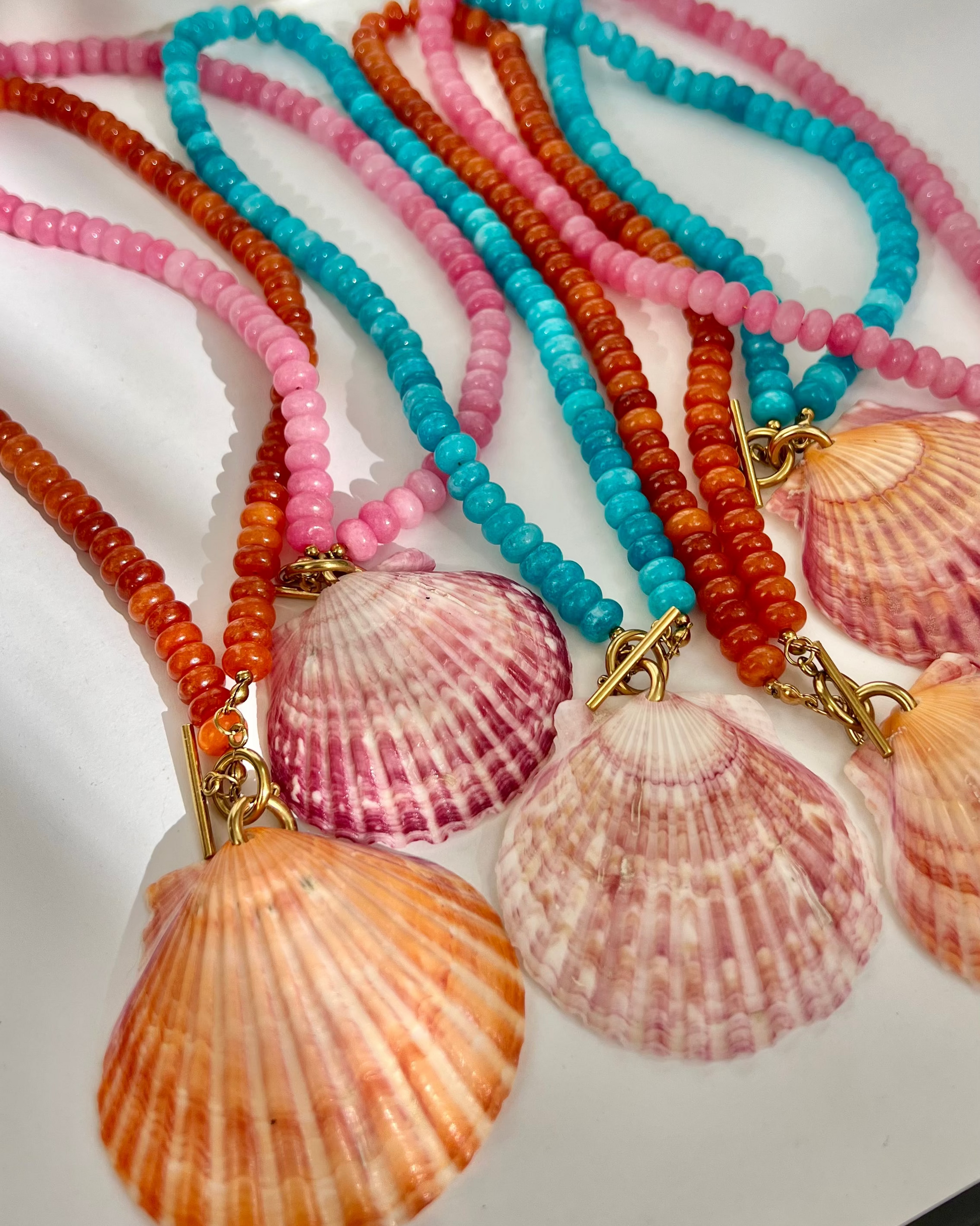 Several beaded shell necklaces arranged on a white surface, each with a large scallop shell pendant and cords in pink, turquoise, orange, and coral tones.