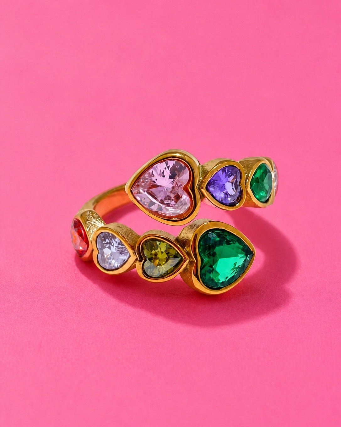 A gold ring with multiple heart-shaped clear gemstones in different colors, including pink, purple, green, and yellow, set against a solid pink background.