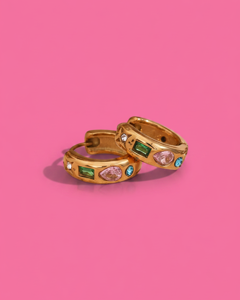 Two gold huggie hoop earrings with colorful gemstones, displayed on a solid pink background.