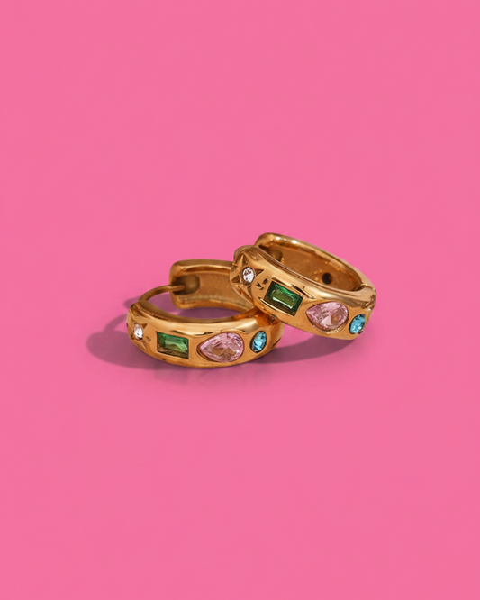 Two gold huggie hoop earrings with colorful gemstones, displayed on a solid pink background.