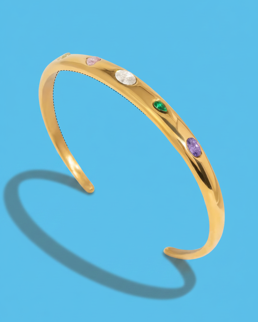 A polished gold cuff bracelet featuring five small, colorful oval gemstones in pink, white, green, and purple, displayed on a solid blue background with a soft shadow.