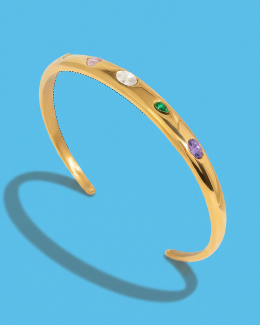 A polished gold cuff bracelet featuring five small, colorful oval gemstones in pink, white, green, and purple, displayed on a solid blue background with a soft shadow.