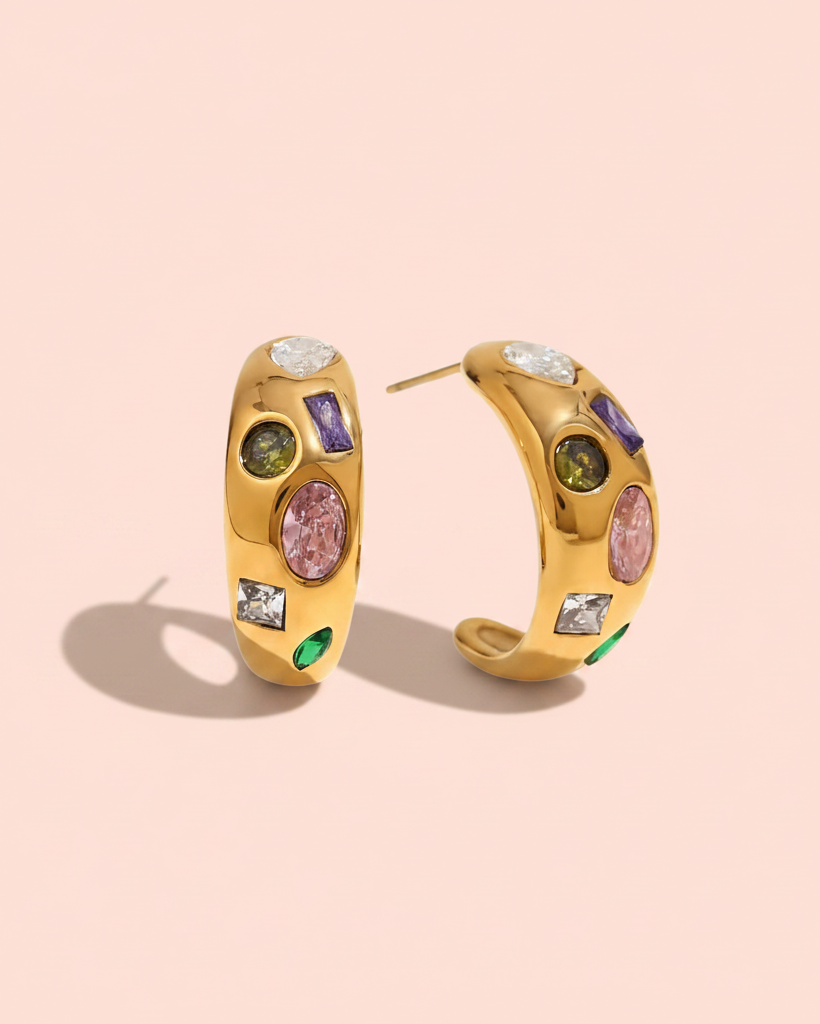 Gold hoop earrings adorned with colorful gemstones, including pink, green, purple, and clear stones, displayed on a soft pink background.